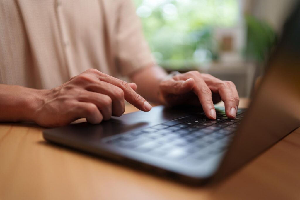 A person sits behind a black laptop at a wooden desk typing on the keyboard.